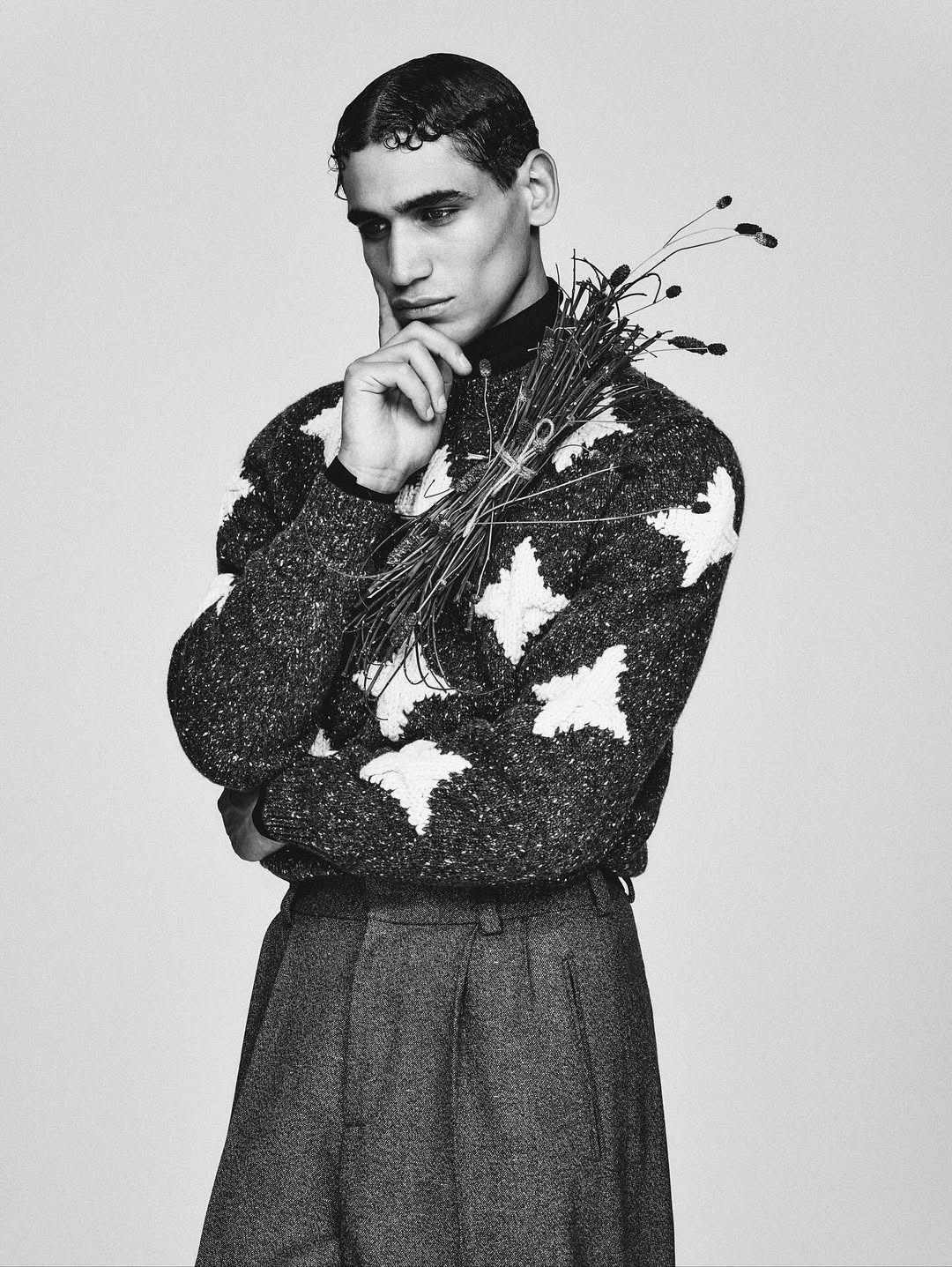 Black and white portrait of a young adult male fashion model in sweater and trousers, holding a bouquet of dried grasses.