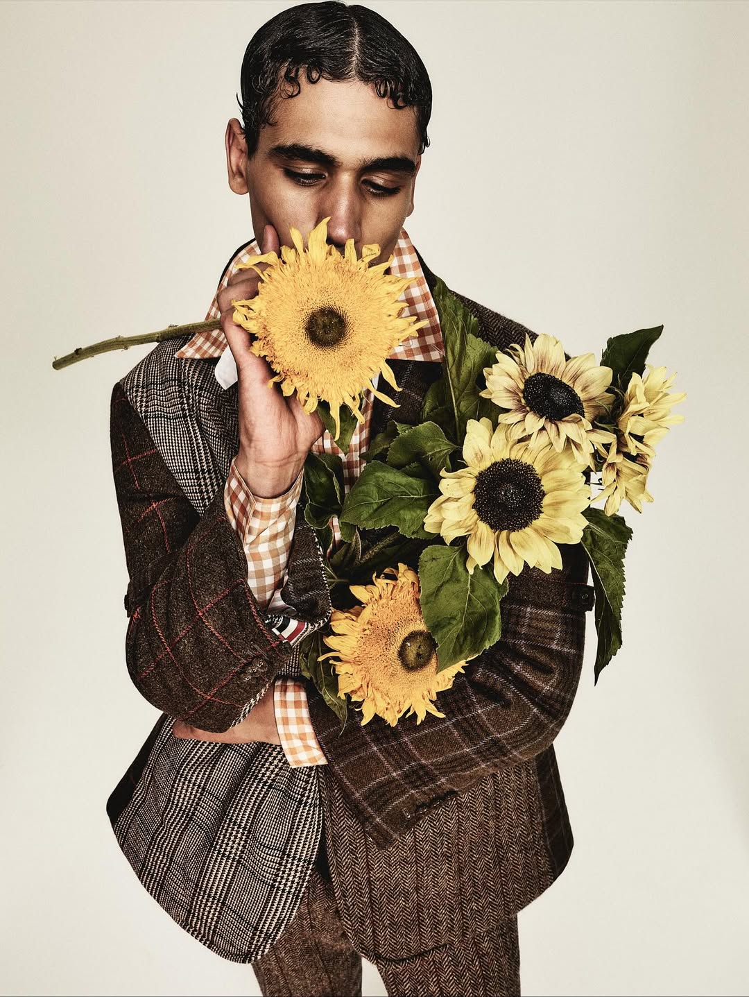 Adult male in plaid suit jacket holding a bouquet of sunflowers in a studio setting, creating a visually striking editorial portrait.