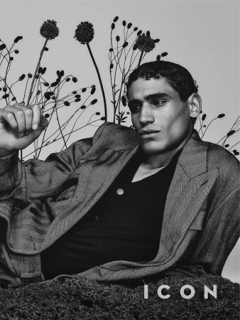 Monochrome portrait of a man in a suit jacket lounging on mossy ground, contrasted with tall, spindly, flowering plants.