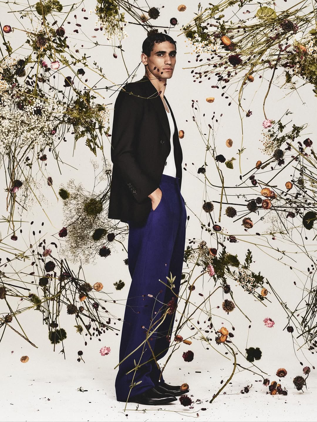 Man in blazer and blue trousers stands among wildflowers in studio setting, fashion editorial image, full-length shot with neutral backdrop.