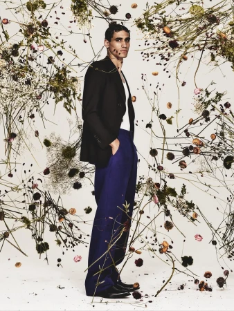Man in blazer and blue trousers stands among wildflowers in studio setting, fashion editorial image, full-length shot with neutral backdrop.
