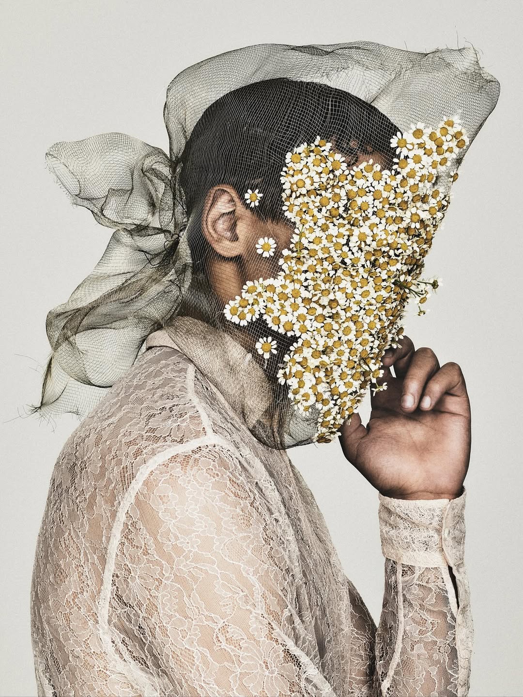 Conceptual portrait of a person wearing lace and mesh with flowers covering their face, set against a light background.