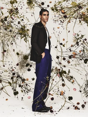 Man standing in dark blazer and blue trousers framed by floating dried flowers against a white background.