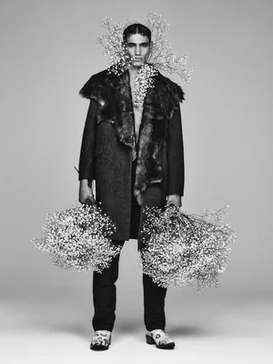 Man wearing a fur-trimmed coat and holding large floral bouquets for ICON Magazine Mena