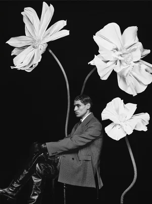 Man seated in a tailored double-breasted blazer and tall leather boots next to giant sculpted flowers against a black backdrop.