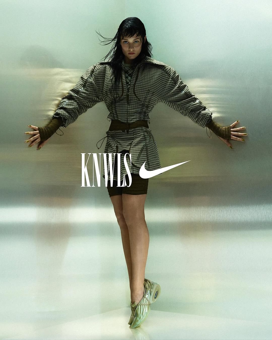 Full shot of a female adult fashion model posing in an earthy-toned outfit with arms outstretched in front of a shiny silver background.
