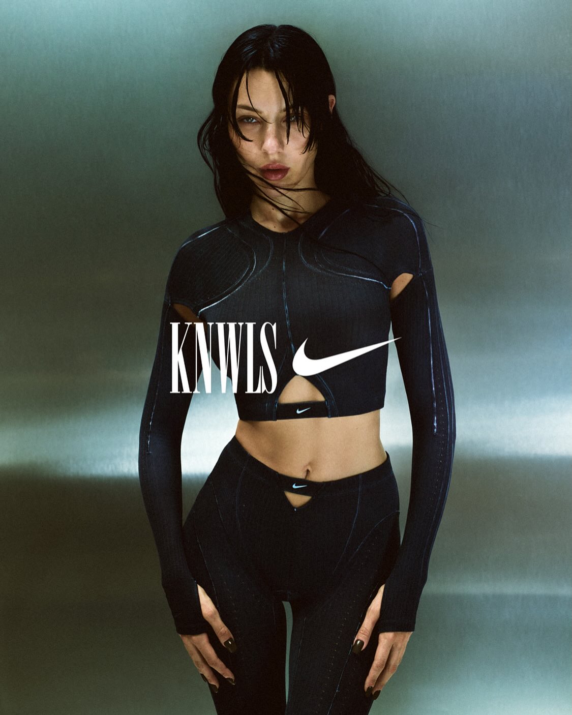 A woman in a black athletic outfit poses against a gray backdrop, featuring a crop top with a Nike logo, matching pants, and the KNWLS logo.