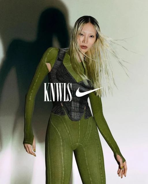 Woman with blonde hair poses in green textured outfit and dark gray vest in studio lighting with KNWLS and Nike brand logos.