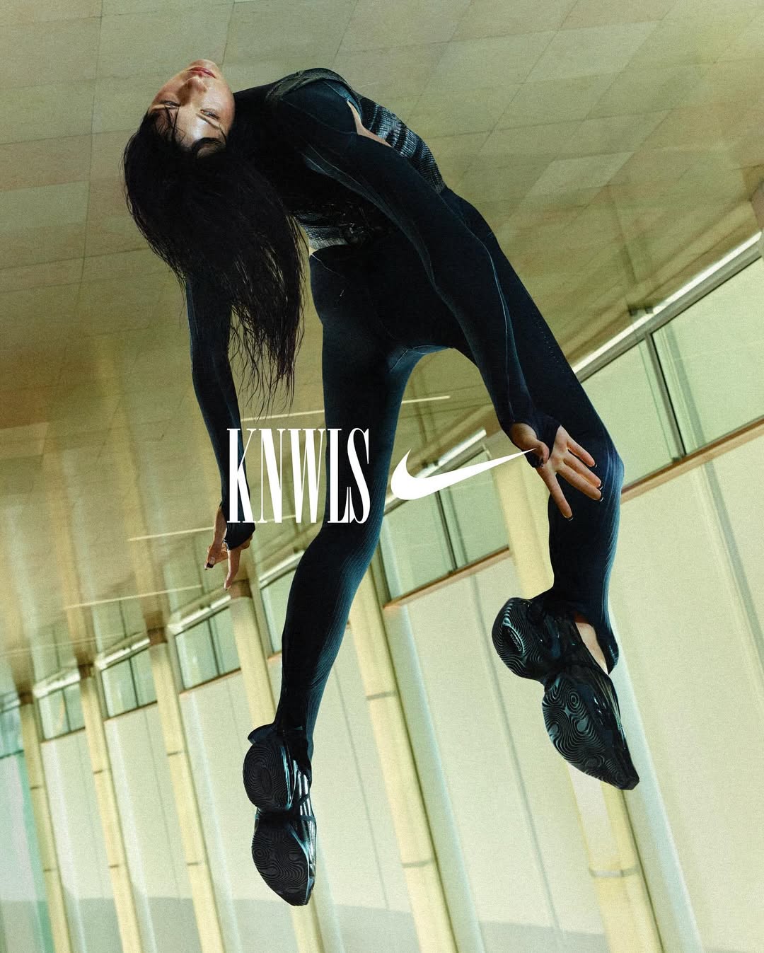 Fashion shot of woman in black activewear and black sneakers in a contorted pose with KNWLS logo and Nike swoosh on a light background.