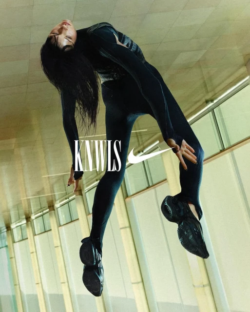 Fashion shot of woman in black activewear and black sneakers in a contorted pose with KNWLS logo and Nike swoosh on a light background.