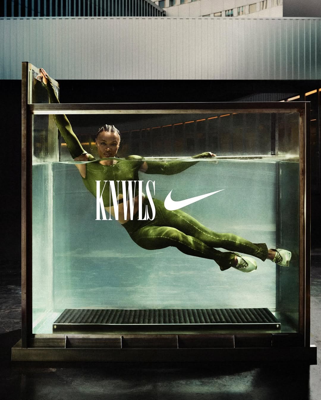 Fashion shot of an adult female in a green bodysuit, partially submerged in a water tank against an architectural backdrop with a Nike logo.