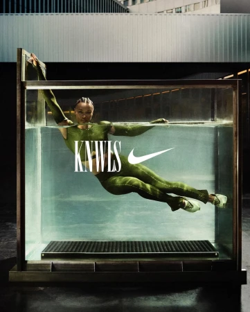 Fashion shot of an adult female in a green bodysuit, partially submerged in a water tank against an architectural backdrop with a Nike logo.