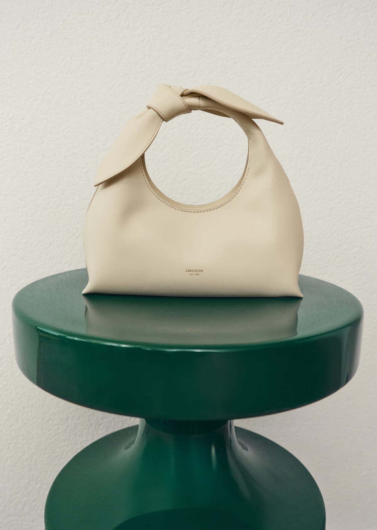 Cream-colored leather top-handle bag with a knotted handle, displayed on a glossy dark green pedestal table against an off-white wall.