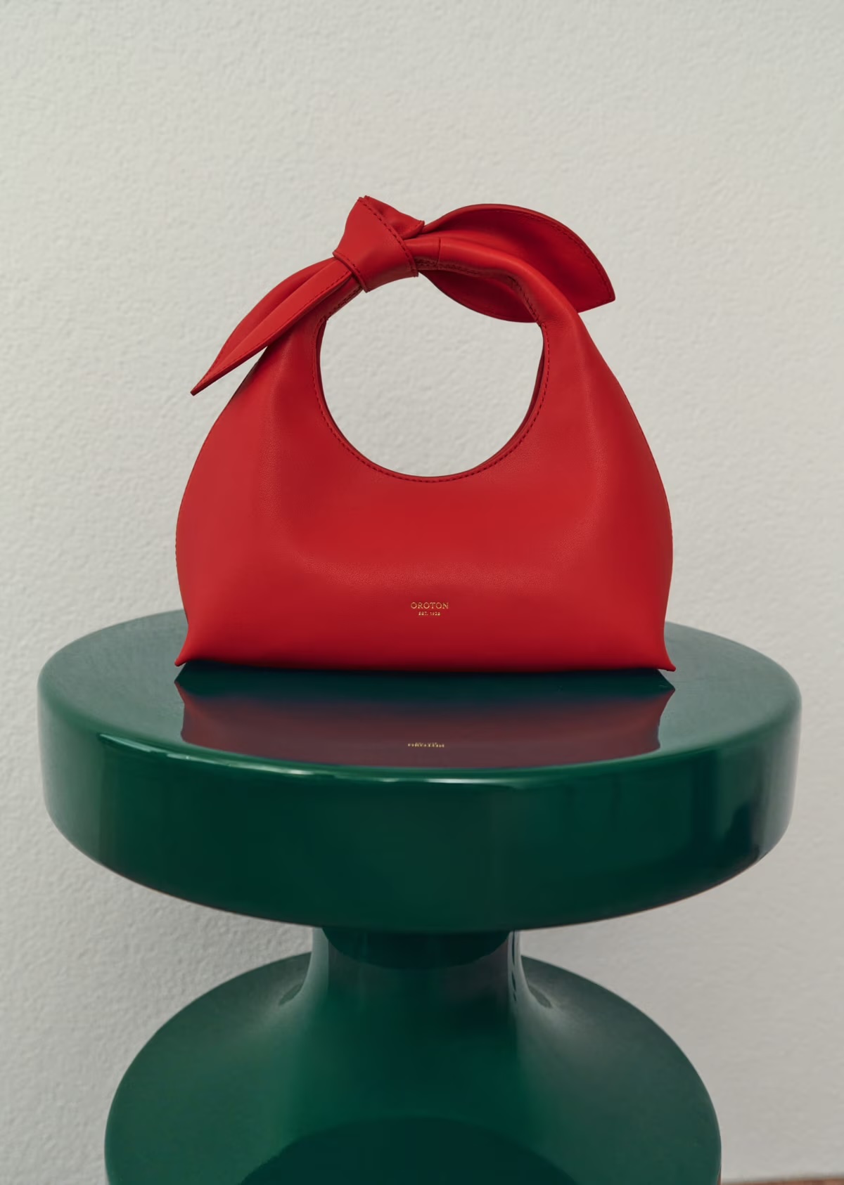 Red leather handbag with bow on a green pedestal stool against a neutral wall, fashion accessory product shot.