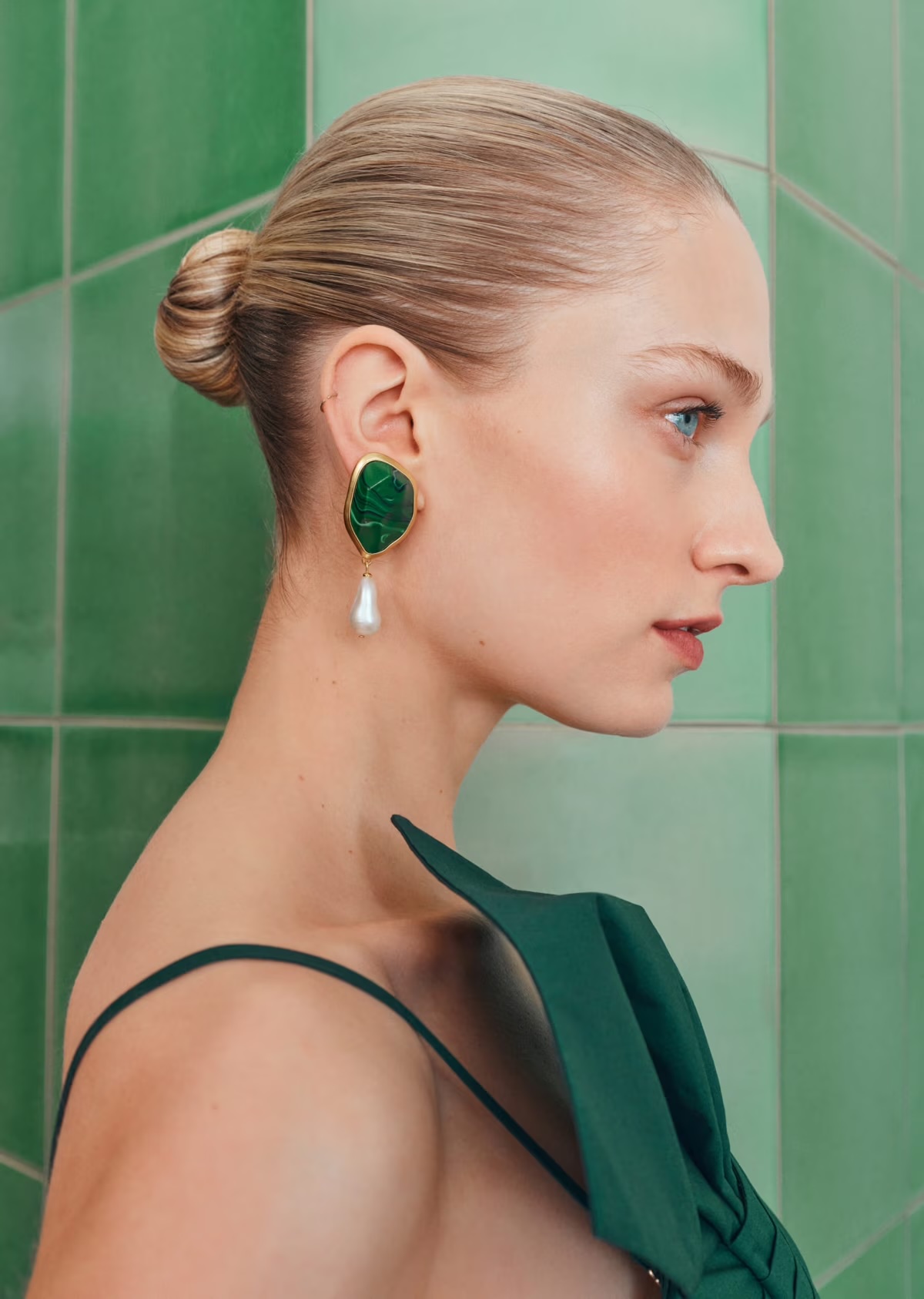 Profile view of a fair-skinned woman with blonde hair in a bun wearing statement green and gold earrings with a pearl drop.