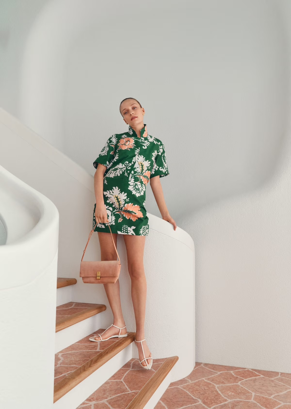 A slender woman poses on indoor stairs wearing a green floral mini dress and carrying a small purse, showcasing a blend of fashion and architecture.