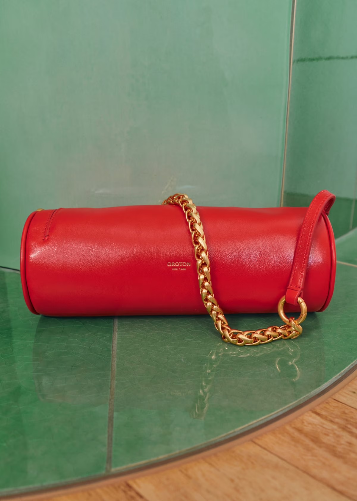 Red cylindrical leather handbag with a gold chain strap on a glossy, patterned green surface, simple background with a brand logo.
