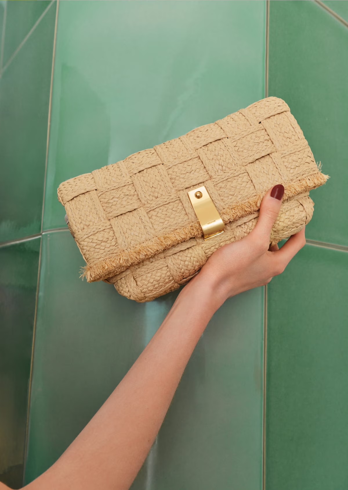 Beige raffia clutch bag with gold clasp, held in a woman's hand, against a green tile background, fashion studio product shot.