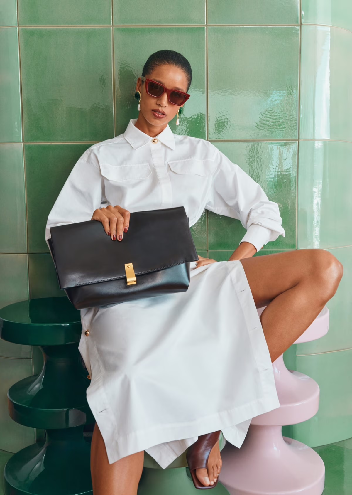 Woman sits on a pink stool wearing a white shirt dress, brown sunglasses, earrings, and sandals while holding a black purse.