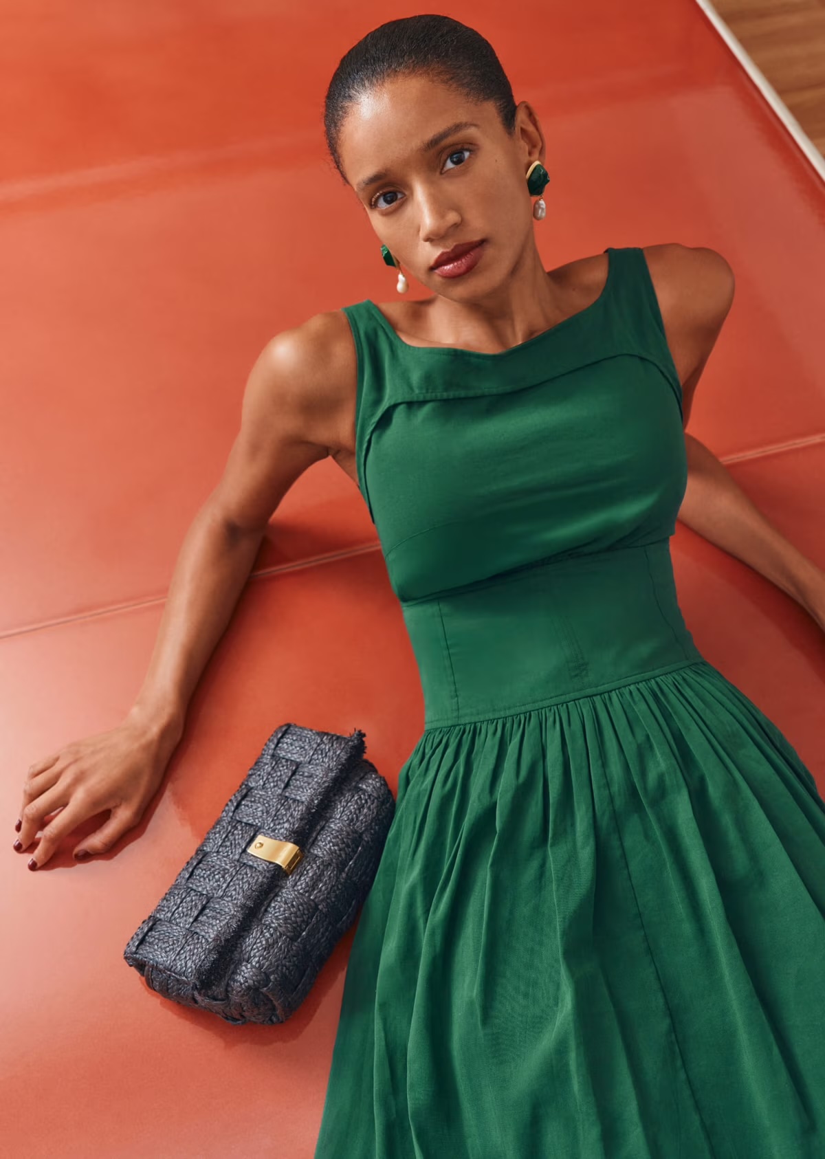 Woman in a green dress with matching earrings and a textured blue clutch poses on a salmon-colored surface in a studio setting.