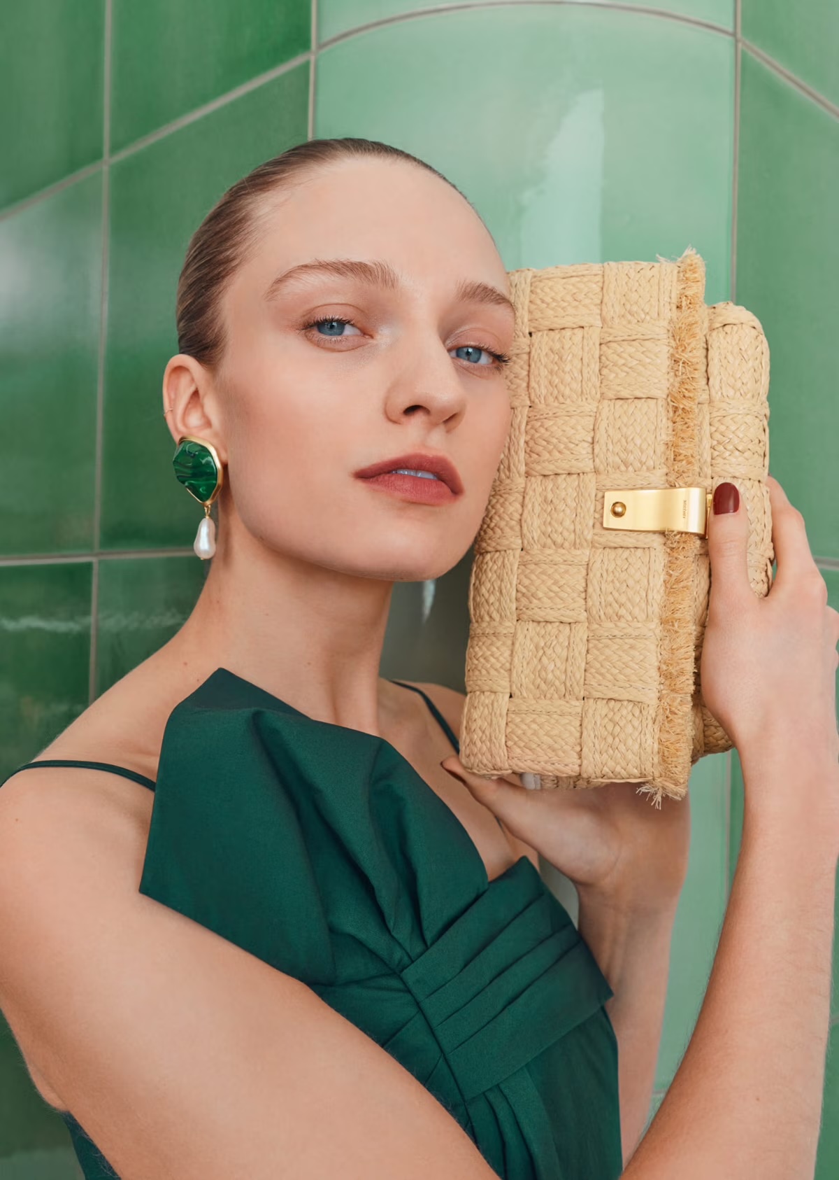 Elegant portrait of a woman holding a woven raffia clutch bag, wearing a green dress and statement earrings against a green tile background.