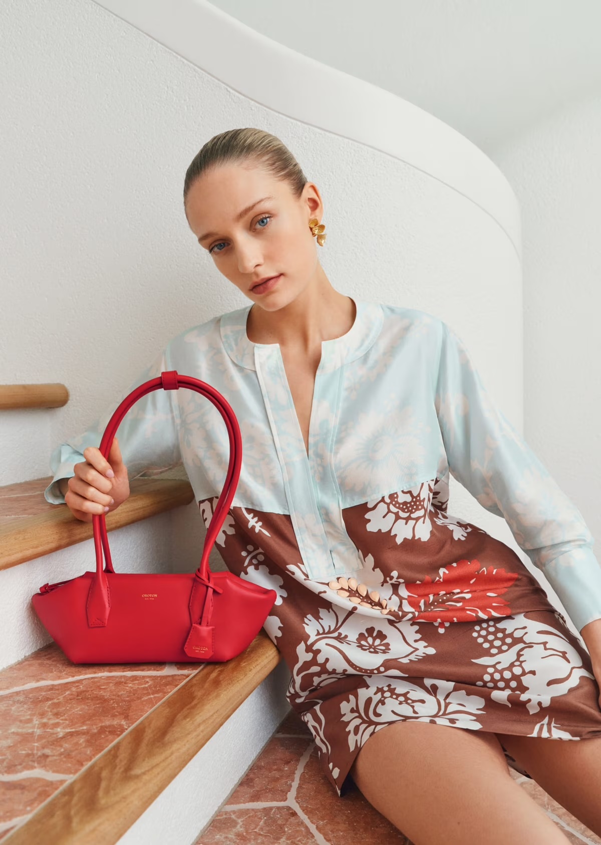 Woman in a dress with floral pattern holding a red handbag poses near a staircase, showcasing fashion and style indoors.
