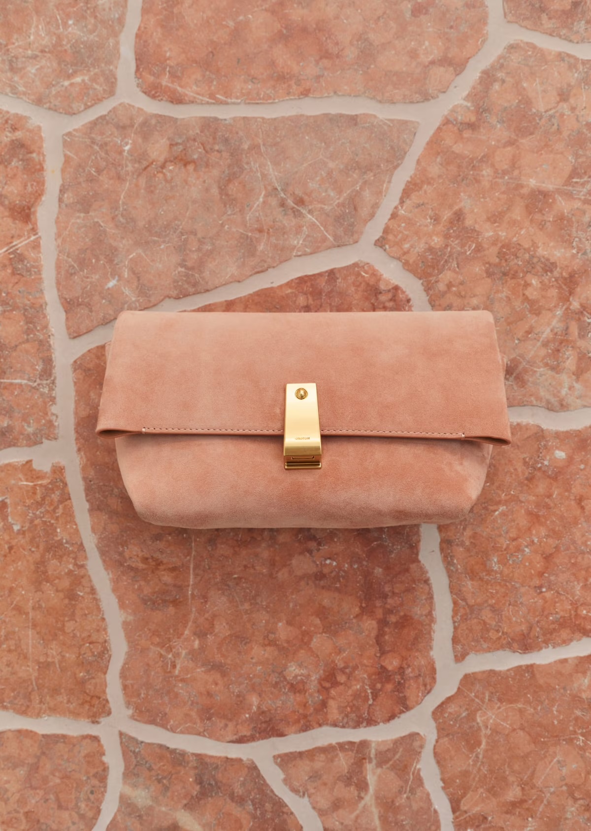 Beige suede clutch bag with gold clasp on a textured orange stone surface, product photography for fashion accessory.