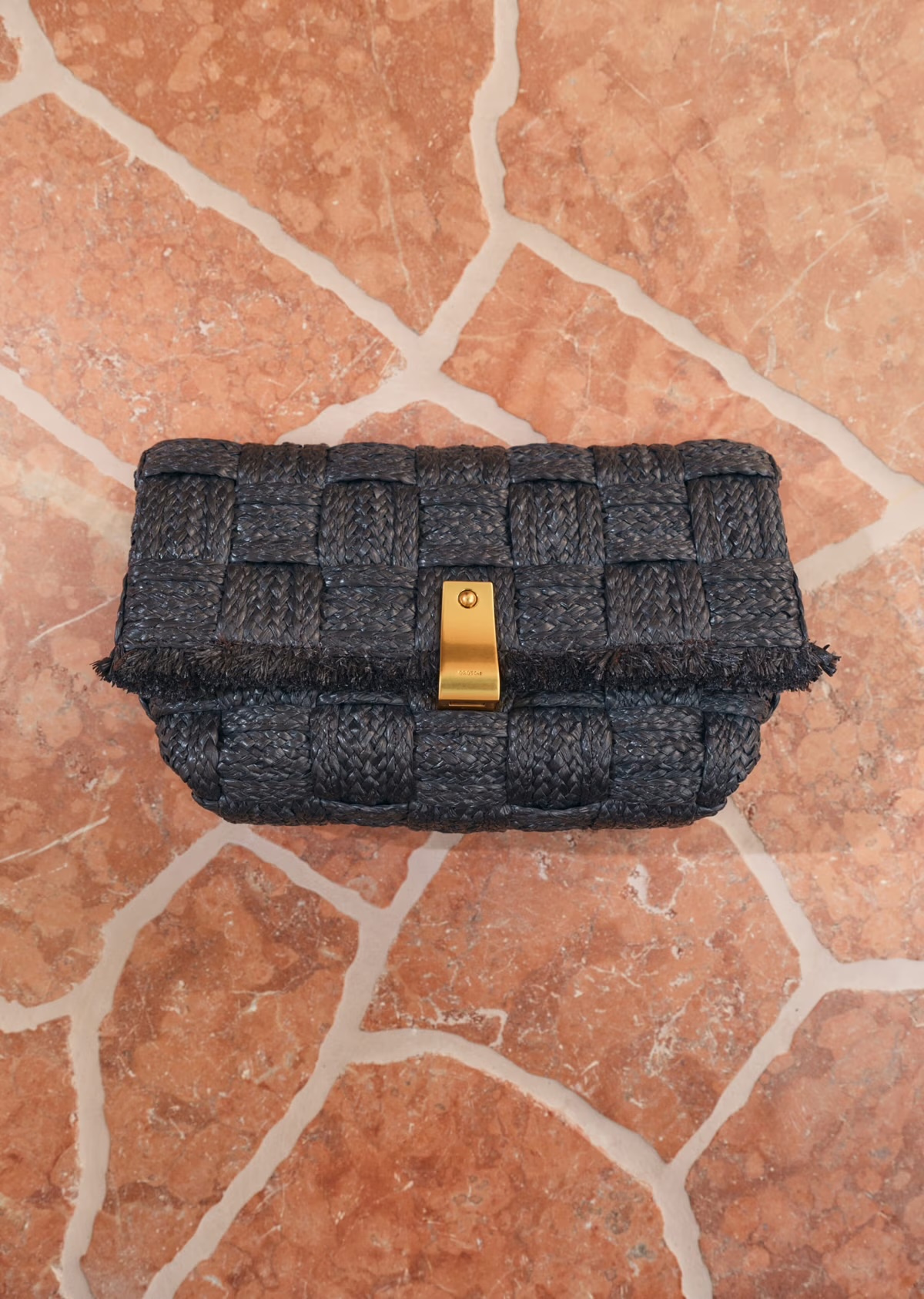 Woven clutch purse in navy with gold clasp, studio shot against a red-brown stone tile background. Fashionable accessory flat lay.