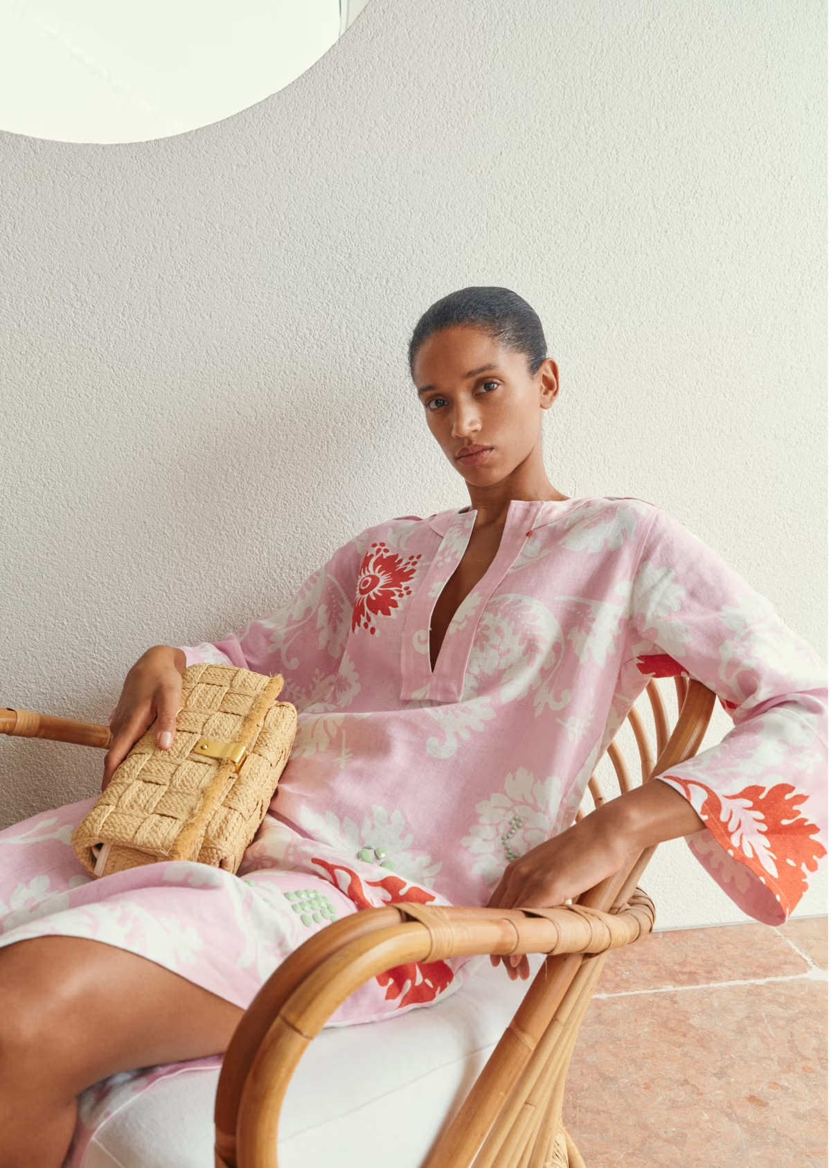 Adult female in a floral dress sitting in a wicker chair, holding a woven clutch. Soft lighting and a neutral-toned wall.