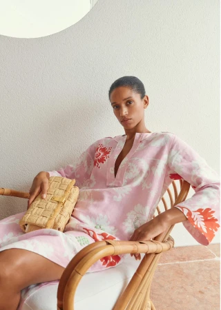 Adult female in a floral dress sitting in a wicker chair, holding a woven clutch. Soft lighting and a neutral-toned wall.