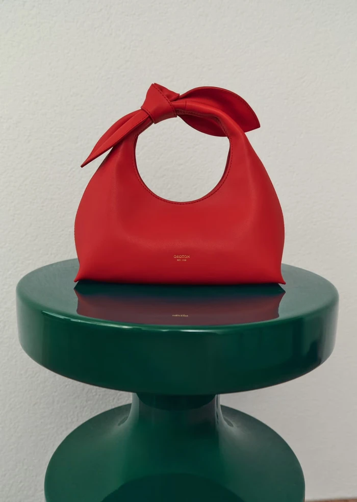 Red leather handbag with knotted handle resting on a glossy green pedestal against a white wall.