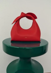 Red leather handbag with knotted handle resting on a glossy green pedestal against a white wall.