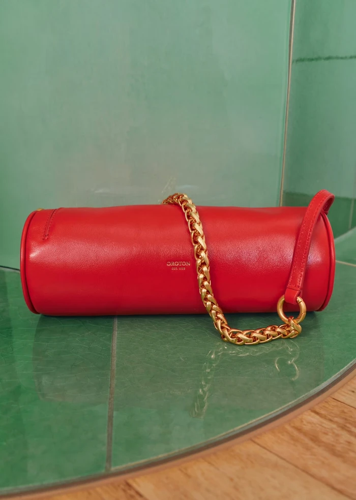 Red cylindrical leather handbag with a thick gold chain strap resting on a green tiled surface.