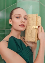 Model in green top holding woven straw clutch bag against glossy green tiled wall