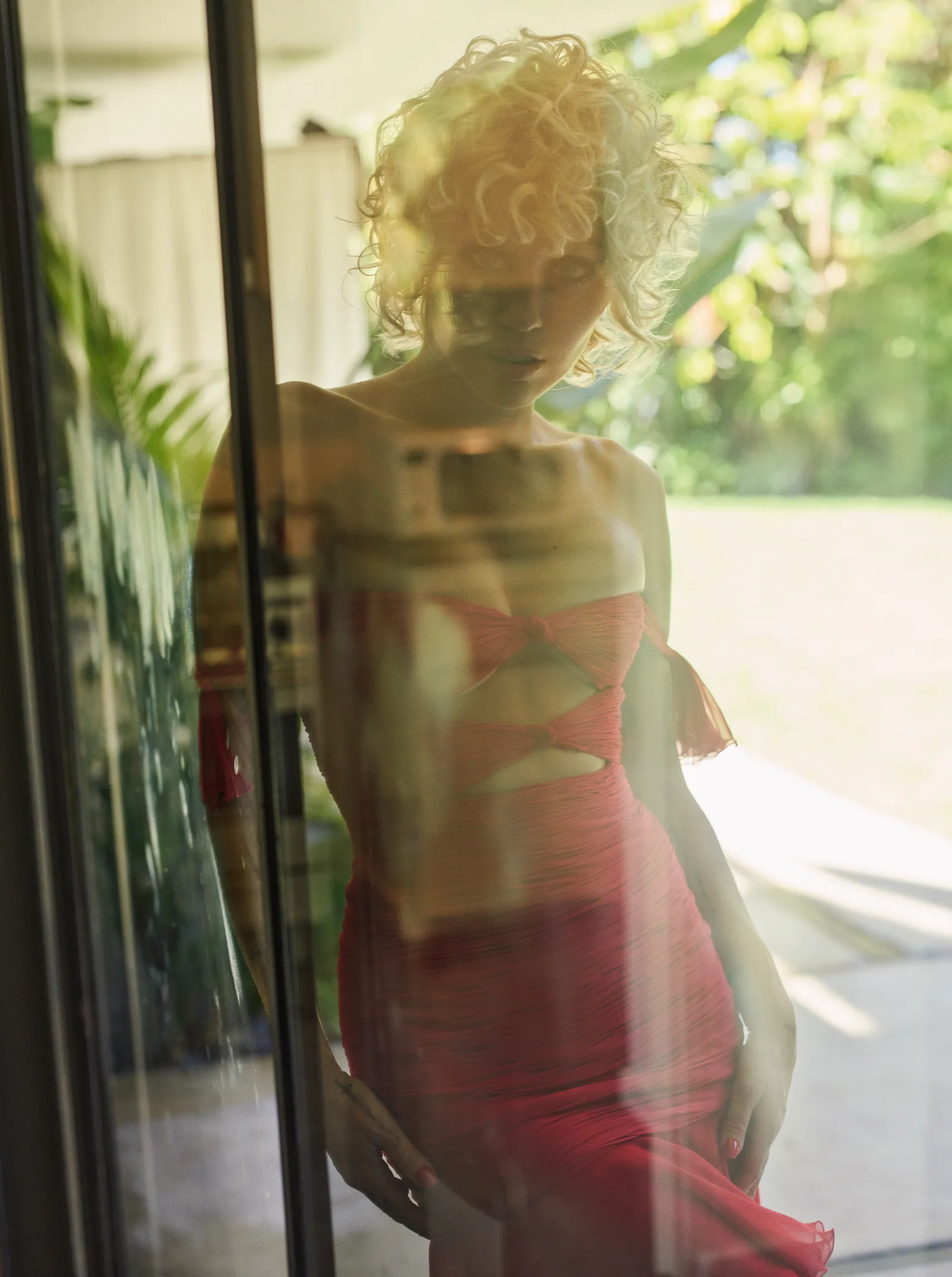 Woman in a red dress poses near a glass door reflecting a green outdoor scene, bathed in soft, natural light. Fashion and lifestyle.