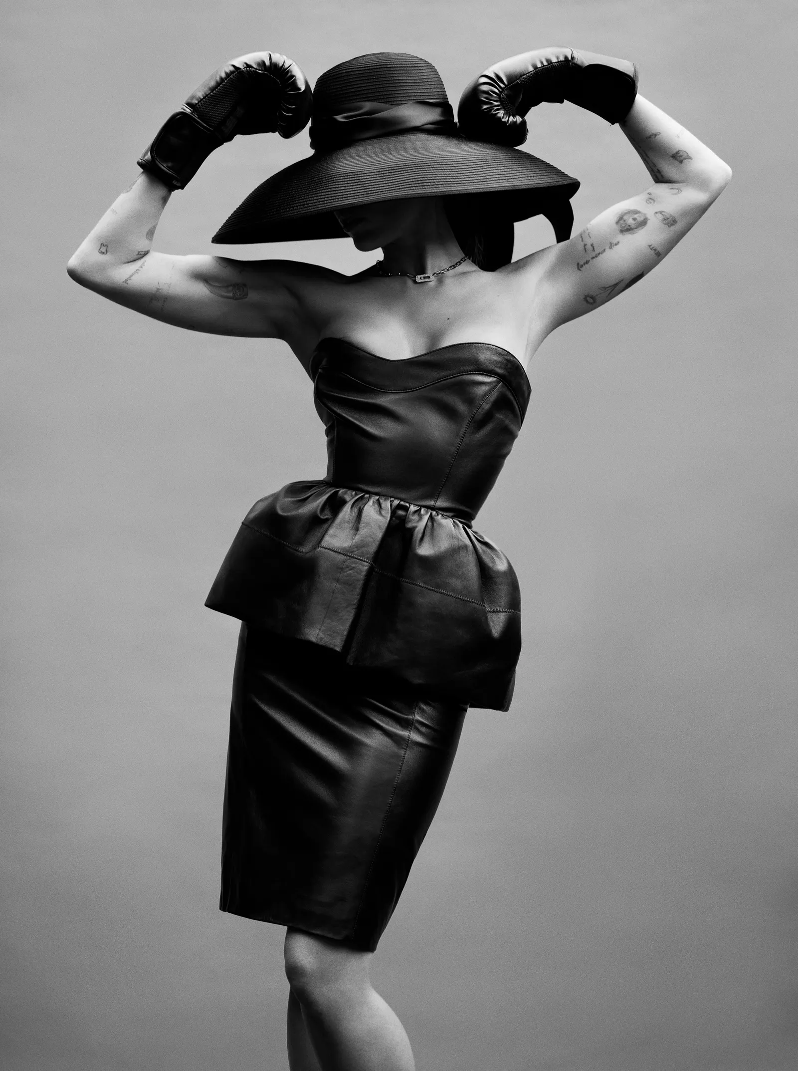 Black and white portrait of a woman in a black hat, leather dress, and boxing gloves posing in a photo studio with a grey background.