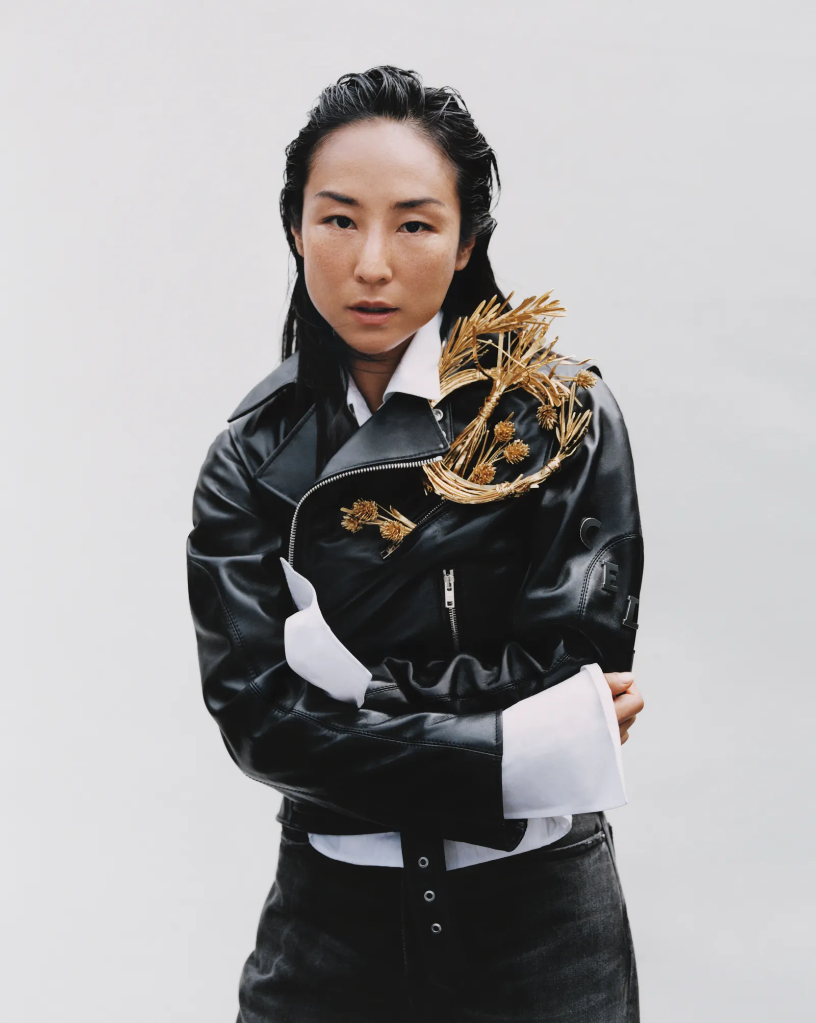 A woman in a black leather jacket with gold embellishments and dark jeans strikes a confident pose against a bright, neutral backdrop.