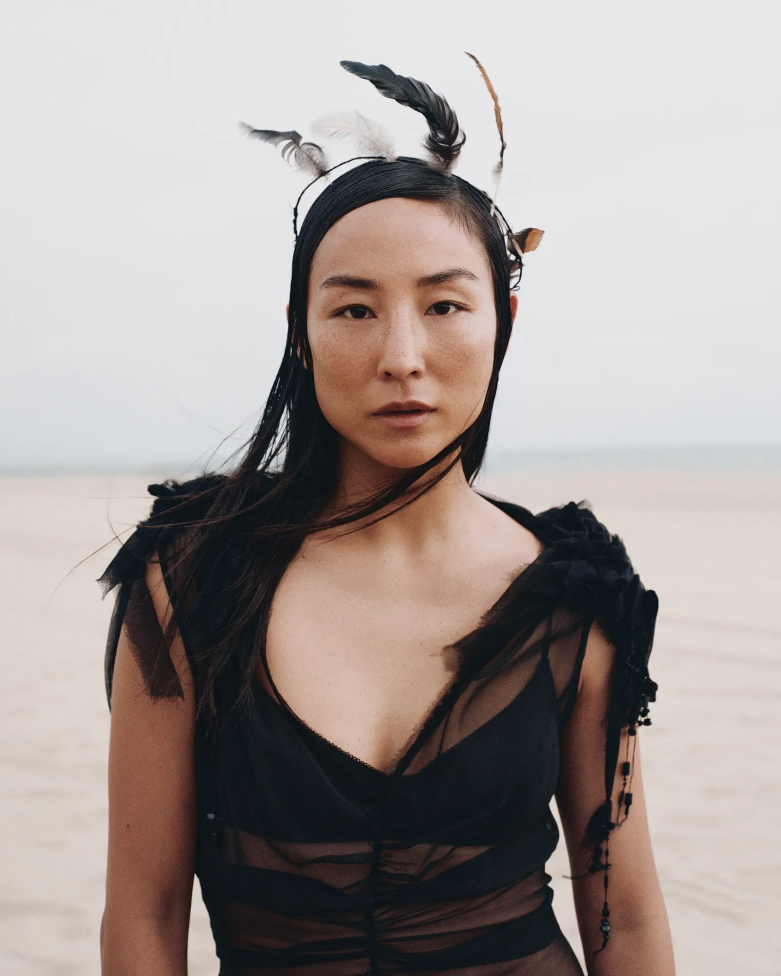 Portrait of an Asian woman with feathered headwear. She wears a sheer black top and stands on a neutral-colored sand beach.