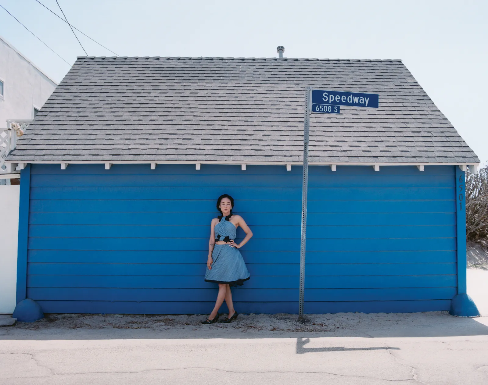 Full shot of an adult woman in a blue dress standing in front of a blue building under a Speedway street sign on a sunny day.
