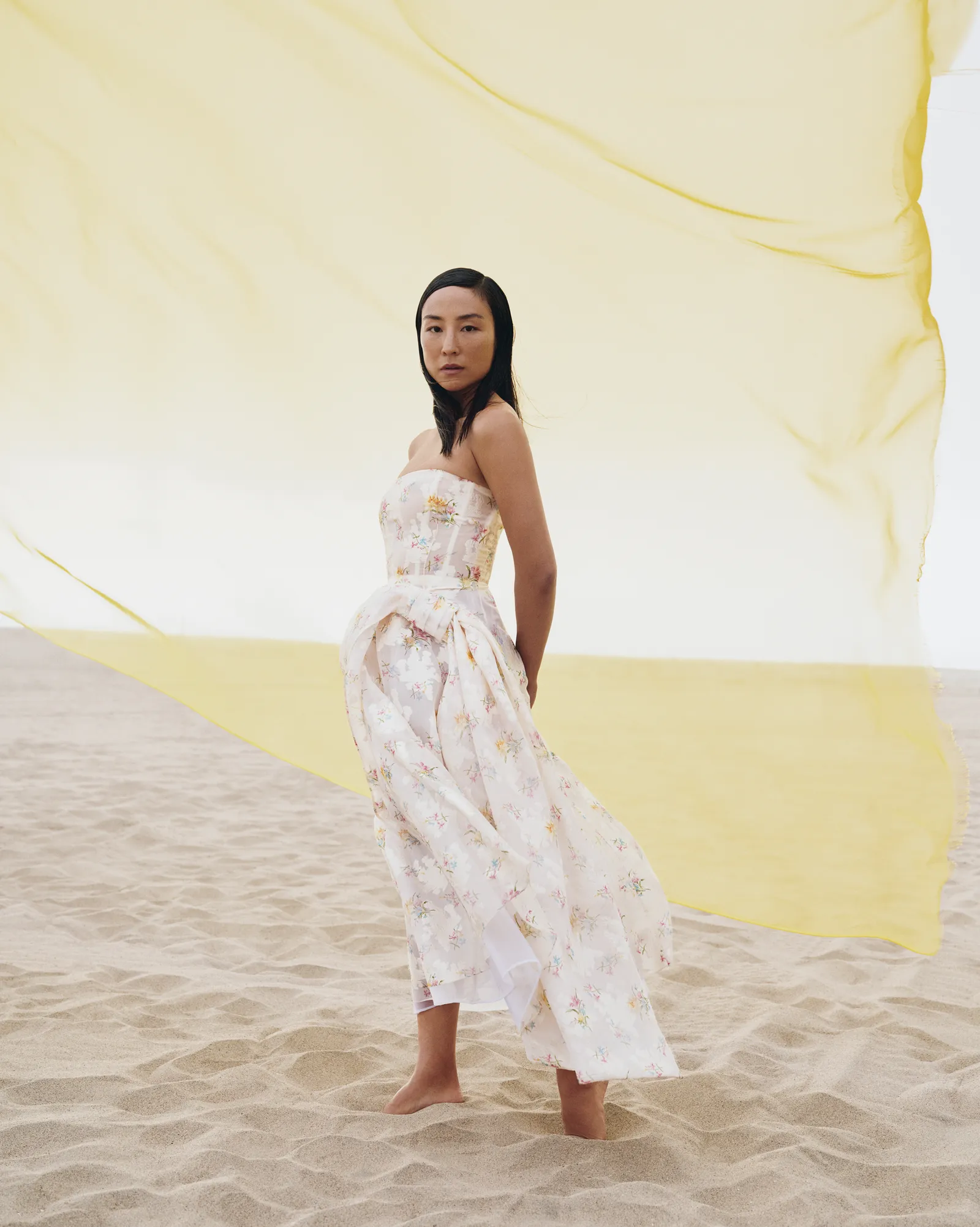 A woman in a floral strapless dress stands on sand in front of a yellow flowing fabric, creating an ethereal and elegant fashion portrait.