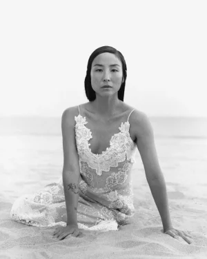 Greta Lee seated on a sandy beach wearing a lace slip dress, photographed by Tyler Mitchell for American Vogue.