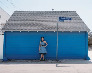 Woman in blue dress standing against a vibrant blue wall below a grey shingled roof and street sign.