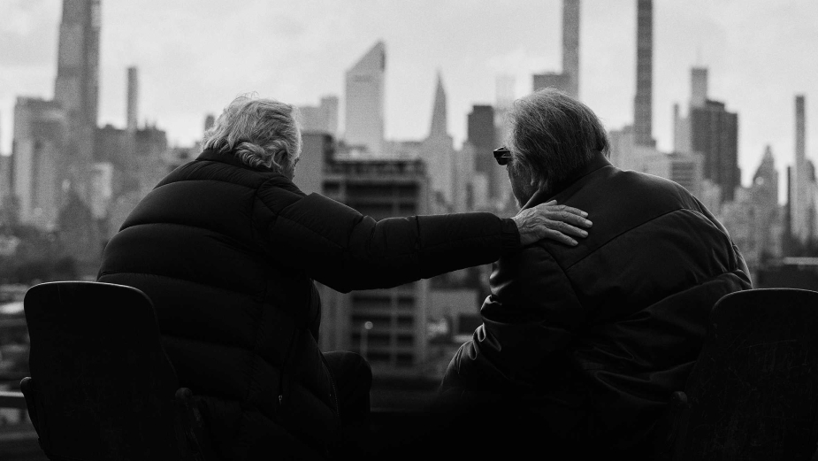 Two elderly people sit together overlooking a cityscape, sharing a quiet moment in an urban setting, with one hand resting on the other's shoulder.