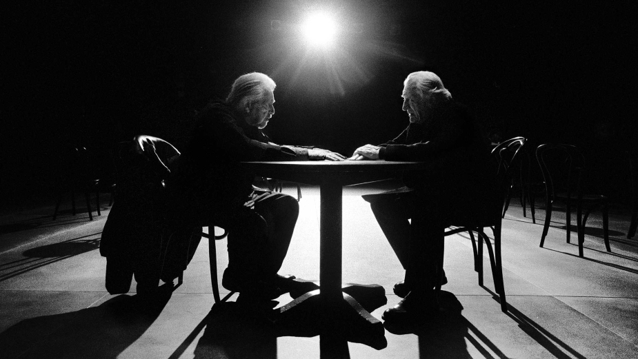 Black and white studio shot featuring two elderly men sitting at a round table, captured with dramatic lighting and minimalist set design.
