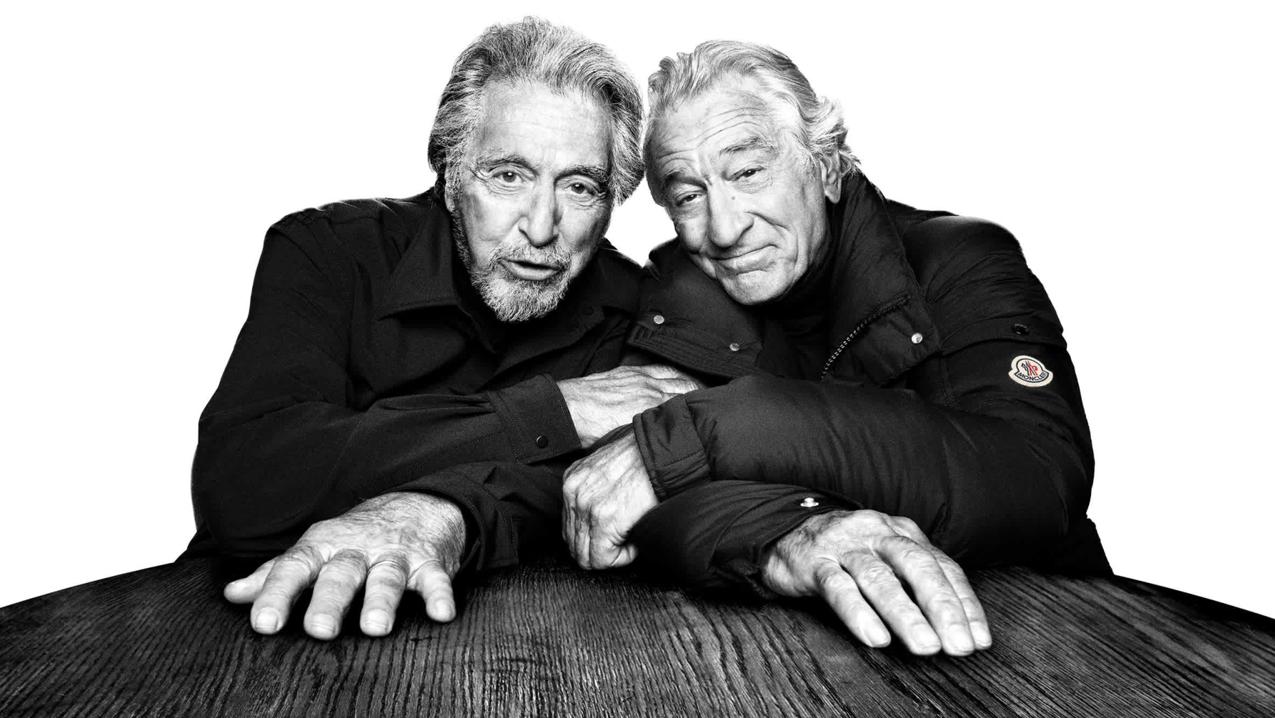 Black and white studio portrait of Al Pacino and Robert De Niro sitting closely at a wooden table looking at the camera.