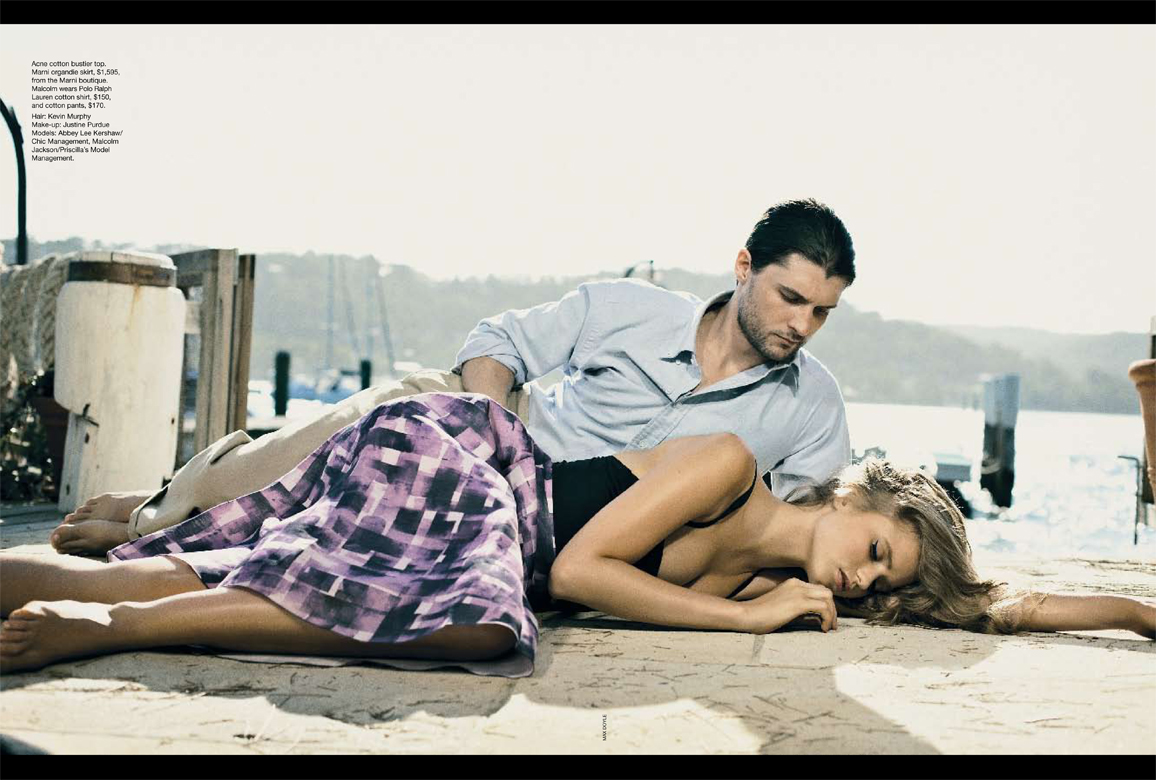 Fashion shot of a couple relaxing outdoors on a pier near a lake, with the woman in a patterned dress and the man in a light blue shirt.