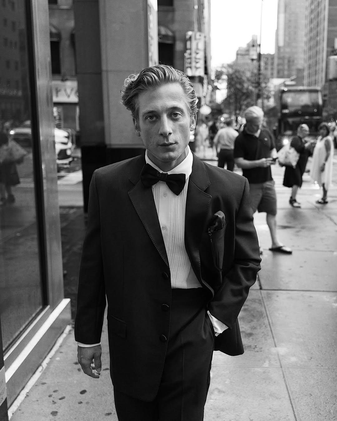 Black and white portrait of a man in a tuxedo on a city sidewalk, surrounded by urban architecture and passersby. Formal wear.