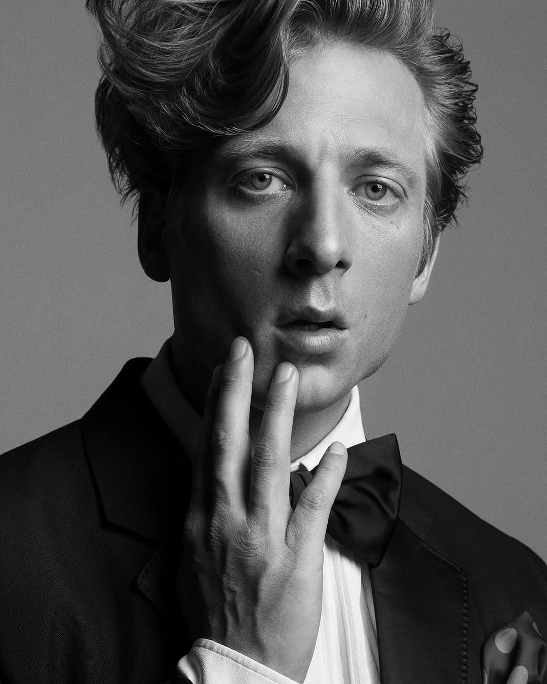 Portrait of an adult male with fair skin, wearing a suit and bow tie, with his hand near his face, set against a grey background.