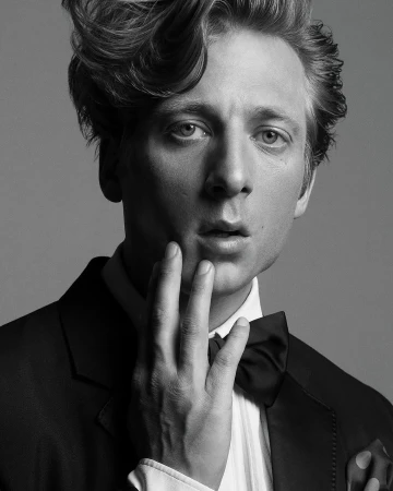 Portrait of an adult male with fair skin, wearing a suit and bow tie, with his hand near his face, set against a grey background.