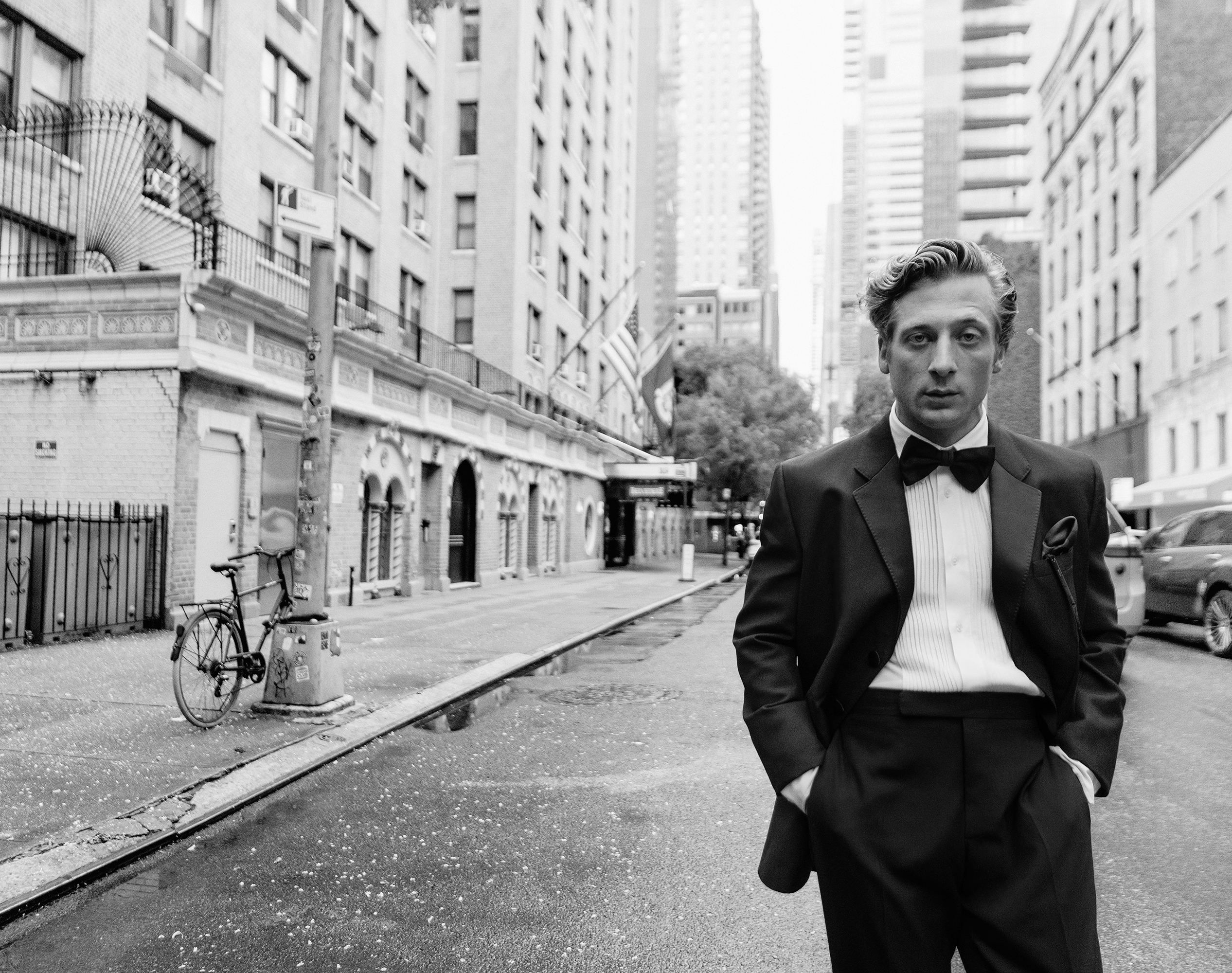 Black-and-white portrait of a man in a tuxedo with bow tie on a city street, featuring architecture, a bicycle, and a car in the background.